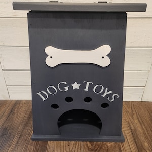 Może przedstawiać: Drewniana skrzynka na zabawki dla psa w kolorze ciemnoszarym z wyciętym białym kością i napisem "DOG * TOYS" białymi literami. Skrzynka ma wycięty ślad łapy i otw&oacute;r do przechowywania zabawek.