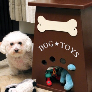 Może przedstawiać: Brązowe drewniane pudełko do przechowywania zabawek dla ps&oacute;w z białym kością i napisem "DOG TOYS" namalowanym z przodu. Pudełko ma wycięcie w kształcie łapy z niebieską i czerwoną zabawką elfa w środku.