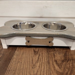 May include: A white and gray wooden dog bowl stand with two stainless steel bowls. The stand has a bone-shaped handle with paw prints.