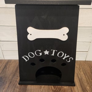 Może przedstawiać: Czarne drewniane pudełko do przechowywania zabawek dla psa z białym kością i napisem "DOG TOYS" z przodu. Pudełko ma wycięcie w kształcie łapy psa z czterema otworami do przechowywania zabawek.