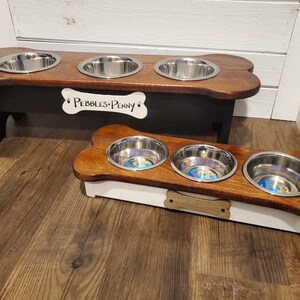 May include: Two wooden dog bowls with bone-shaped bases. The larger bowl is black with a white bone-shaped plaque that reads "Pebbles & Penny". The smaller bowl is white with a brown bone-shaped handle.