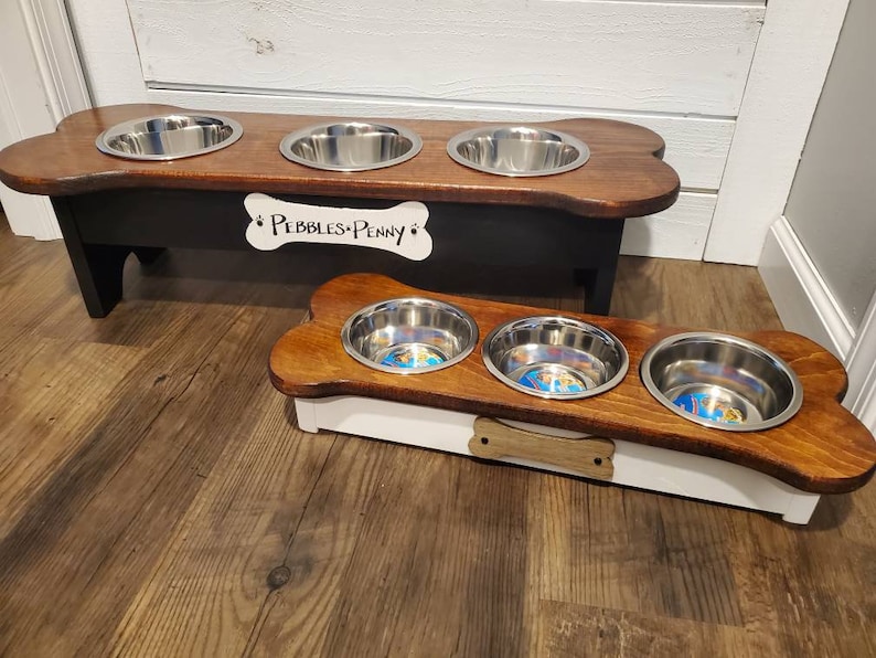 May include: Two wooden dog bowls with bone-shaped bases. The larger bowl is black with a white bone-shaped plaque that reads "Pebbles & Penny". The smaller bowl is white with a brown bone-shaped handle.