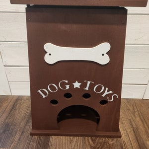 Może przedstawiać: Brązowe drewniane pudełko do przechowywania zabawek dla ps&oacute;w z białym kością i napisem "DOG TOYS" z przodu. Pudełko ma mały otw&oacute;r z przodu, aby psy mogły dostać się do swoich zabawek.