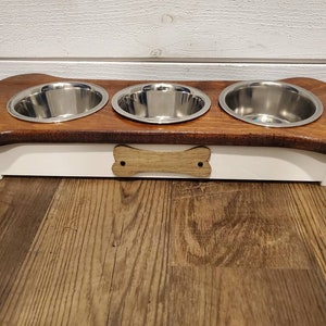 May include: A wooden dog bowl stand with three stainless steel bowls. The stand is shaped like a bone and has a white base with a wooden bone design.
