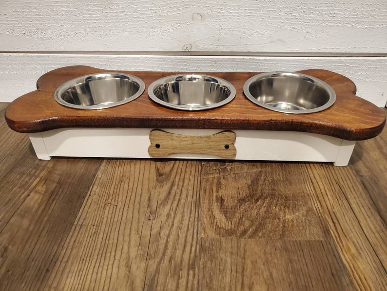 May include: A wooden dog bowl stand with three stainless steel bowls. The stand is shaped like a bone and has a white base with a wooden bone design.