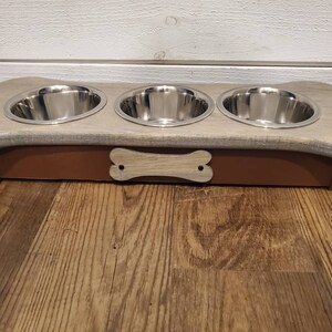 May include: A wooden dog bowl stand shaped like a bone with three stainless steel bowls. The stand is painted a light gray with a brown base and a bone-shaped cutout on the front.