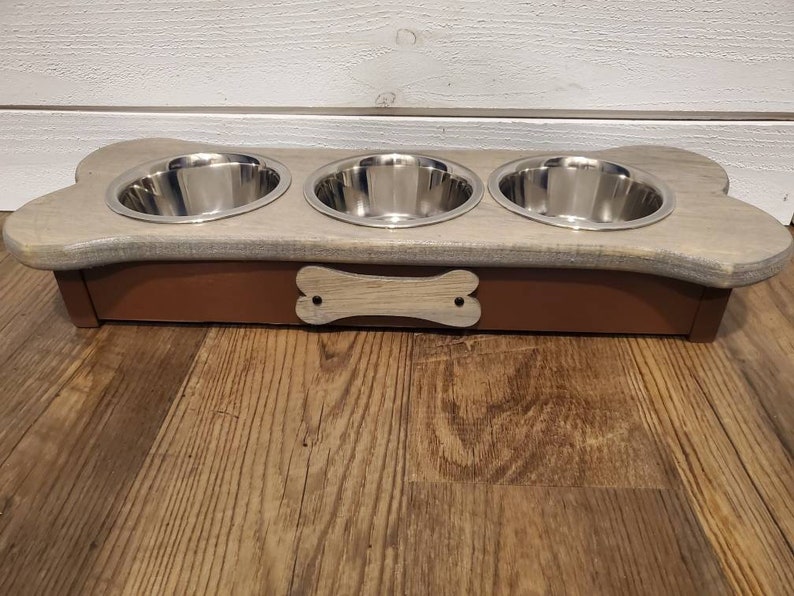 May include: A wooden dog bowl stand shaped like a bone with three stainless steel bowls. The stand is painted a light gray with a brown base and a bone-shaped cutout on the front.