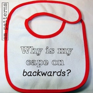 May include: White baby bib with a red border. The bib has the text "Why is my cape on backwards?" printed on it.
