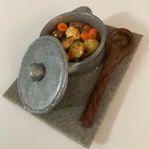 May include: A silver pot with a lid filled with a stew of potatoes, carrots, peas, and meat. The pot is sitting on a gray stone surface with a wooden spoon next to it.