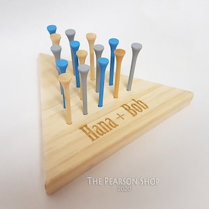 May include: A wooden peg game with a triangular base and colorful pegs. The pegs are in shades of blue, gray, and natural wood. The base is engraved with the names "Hana + Bob". The Pearson Shop 2020 is printed at the bottom.