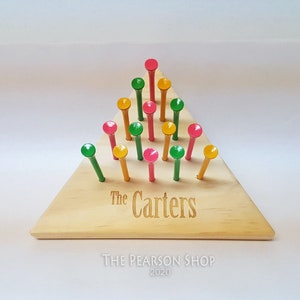 May include: Wooden peg game with a triangular base. The base is light-colored wood, engraved with "The Carters." Colorful pegs in red, green, and yellow are arranged in a triangular pattern. The Pearson Shop 2020 is printed below.