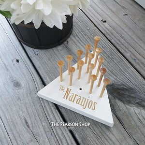 May include: A white wooden peg game with the text "The Naranjos" in gold lettering. The game has wooden pegs and is set on a weathered wooden surface. A bouquet of white flowers is in the background.
