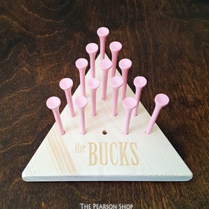 May include: A wooden peg game with a triangular base and pink pegs. The base is light-colored wood with the engraved text "the BUCKS". The game is set against a dark wood background.