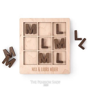 May include: Wooden game board with a grid of nine squares. Each square has a letter 'L' or 'M' printed on it. The board is personalized with the text "Max & Laura Weber" and "The Pearson Shop 2021".