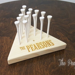 May include: A wooden golf tee holder in a triangular shape, with the text "THE PEARSONS" engraved on the front. The holder contains multiple white golf tees. The background is a dark wood surface.