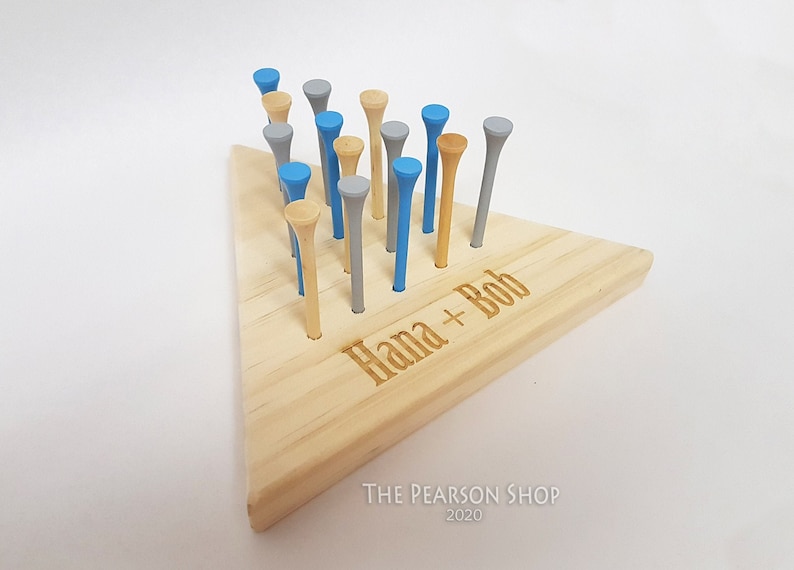 May include: A wooden peg game with a triangular base and colorful pegs. The pegs are in shades of blue, gray, and natural wood. The base is engraved with the names "Hana + Bob". The Pearson Shop 2020 is printed at the bottom.
