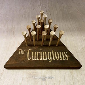 May include: A wooden golf tee holder in a triangular shape, stained dark brown. The holder has the engraved text "The Curingtons" and holds multiple wooden golf tees. The Pearson Shop 2026 is printed at the bottom.