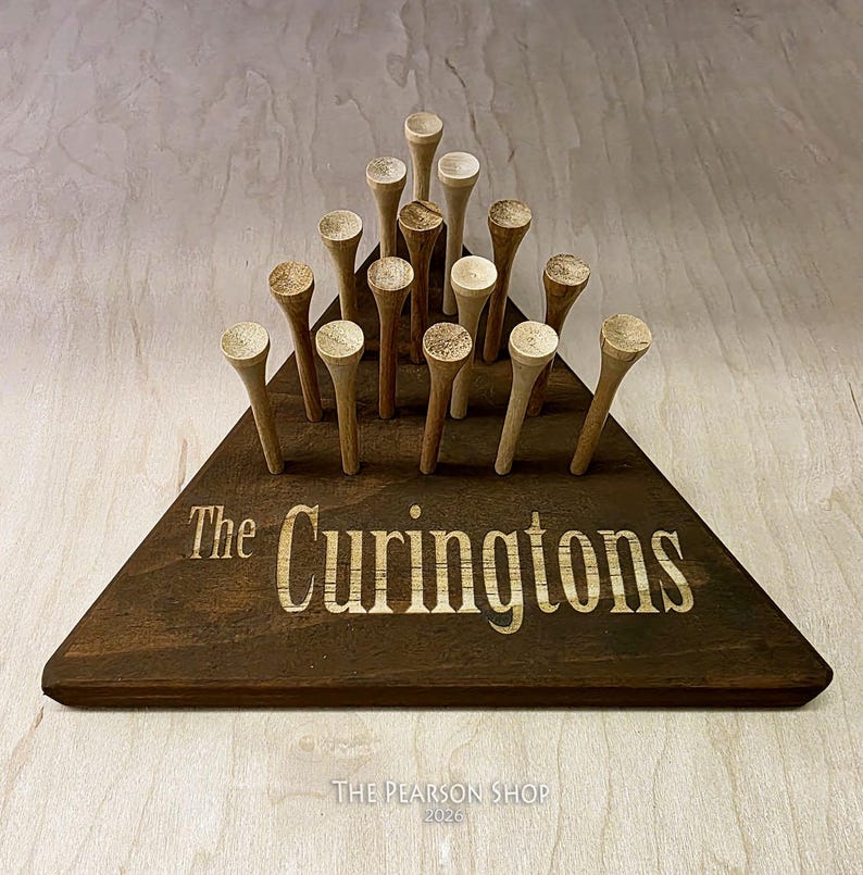 May include: A wooden golf tee holder in a triangular shape, stained dark brown. The holder has the engraved text "The Curingtons" and holds multiple wooden golf tees. The Pearson Shop 2026 is printed at the bottom.