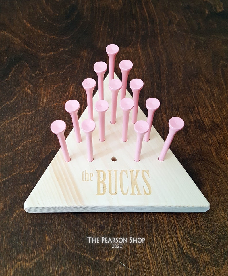 May include: A wooden peg game with a triangular base and pink pegs. The base is light-colored wood with the engraved text "the BUCKS". The game is set against a dark wood background.