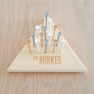 May include: A wooden triangle-shaped game board with golf tees. The board is light-colored wood, with the name "THE BURKES" engraved on it. The golf tees are white, gray, and natural wood-colored. The text "THE PEARSON SHOP 2020" is below the board.
