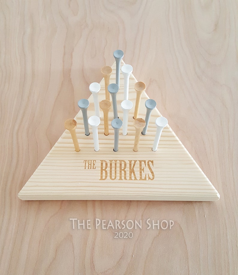 May include: A wooden triangle-shaped game board with golf tees. The board is light-colored wood, with the name "THE BURKES" engraved on it. The golf tees are white, gray, and natural wood-colored. The text "THE PEARSON SHOP 2020" is below the board.