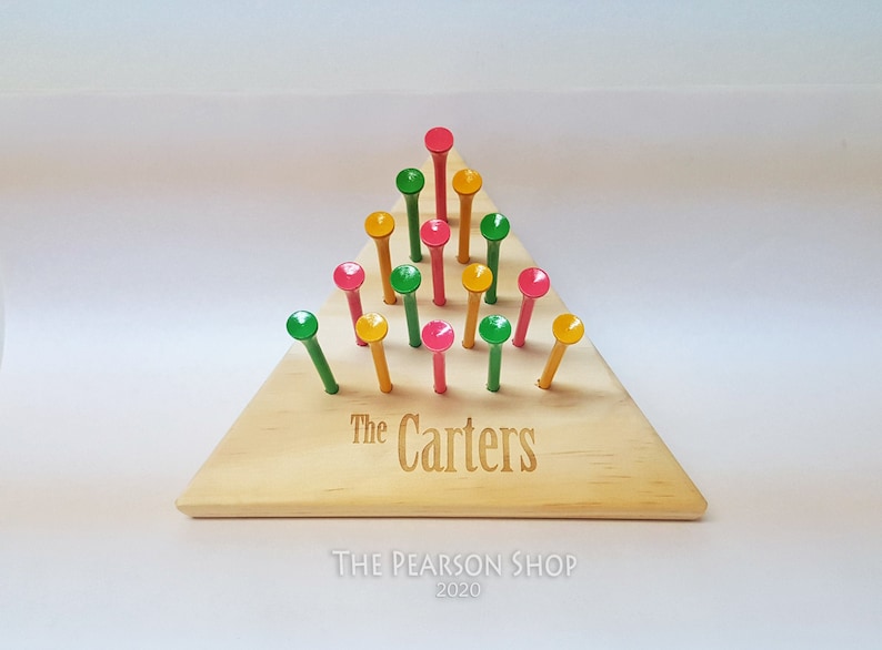 May include: Wooden peg game with a triangular base. The base is light-colored wood, engraved with "The Carters." Colorful pegs in red, green, and yellow are arranged in a triangular pattern. The Pearson Shop 2020 is printed below.