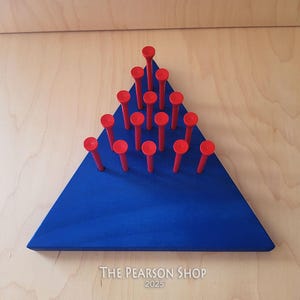 May include: A blue wooden triangle with red pegs arranged in a triangular pattern. The pegs are evenly spaced and have a rounded top. The wooden base has the text "THE PEARSON SHOP 2025" printed on it.