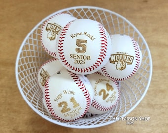 May include: A white wire basket filled with personalized baseballs. Each baseball has red stitching and features a name, number, and the text "SENIOR 2025". Some baseballs also display a wolf logo.