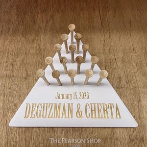 May include: A white triangular wooden board with wooden pegs arranged in a pyramid shape. The board is engraved with the date "January 15, 2026" and the names "DEGUZMAN & CHERTA" in gold lettering. The Pearson Shop 2026 is printed at the bottom.