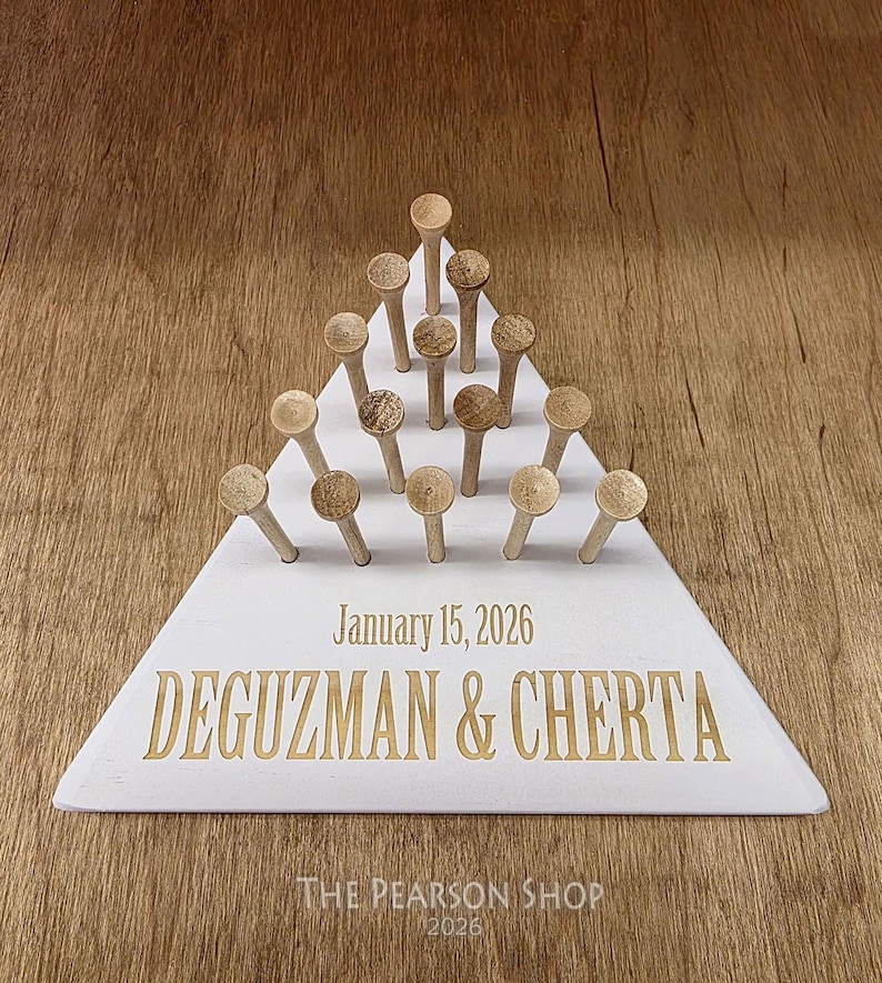 May include: A white triangular wooden board with wooden pegs arranged in a pyramid shape. The board is engraved with the date "January 15, 2026" and the names "DEGUZMAN & CHERTA" in gold lettering. The Pearson Shop 2026 is printed at the bottom.
