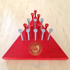 May include: A red wooden peg game with a triangular base. The base features a school emblem and the text "DEERFIELD HIGH SCHOOL WARRIORS". The game includes red and gray pegs. The text "The Pearson Shop 2026" is at the bottom.