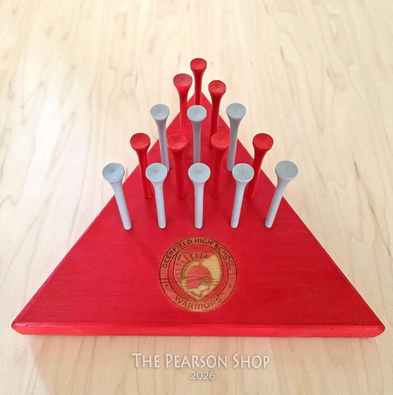 May include: A red wooden peg game with a triangular base. The base features a school emblem and the text "DEERFIELD HIGH SCHOOL WARRIORS". The game includes red and gray pegs. The text "The Pearson Shop 2026" is at the bottom.