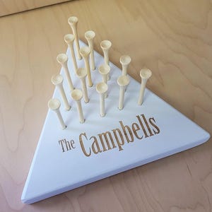May include: A white, triangular wooden game board with wooden pegs. The board is personalized with the text "The Campbells" in a gold color. The game board is on a light wood surface.