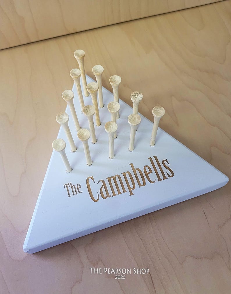 May include: A white, triangular wooden game board with wooden pegs. The board is personalized with the text "The Campbells" in a gold color. The game board is on a light wood surface.