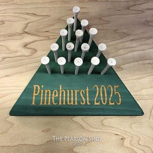 May include: A green wooden golf tee holder in a triangular shape, with the words "Pinehurst 2025" in gold lettering. The holder contains 15 white golf tees. The text "The Pearson Shop 2026" is at the bottom.