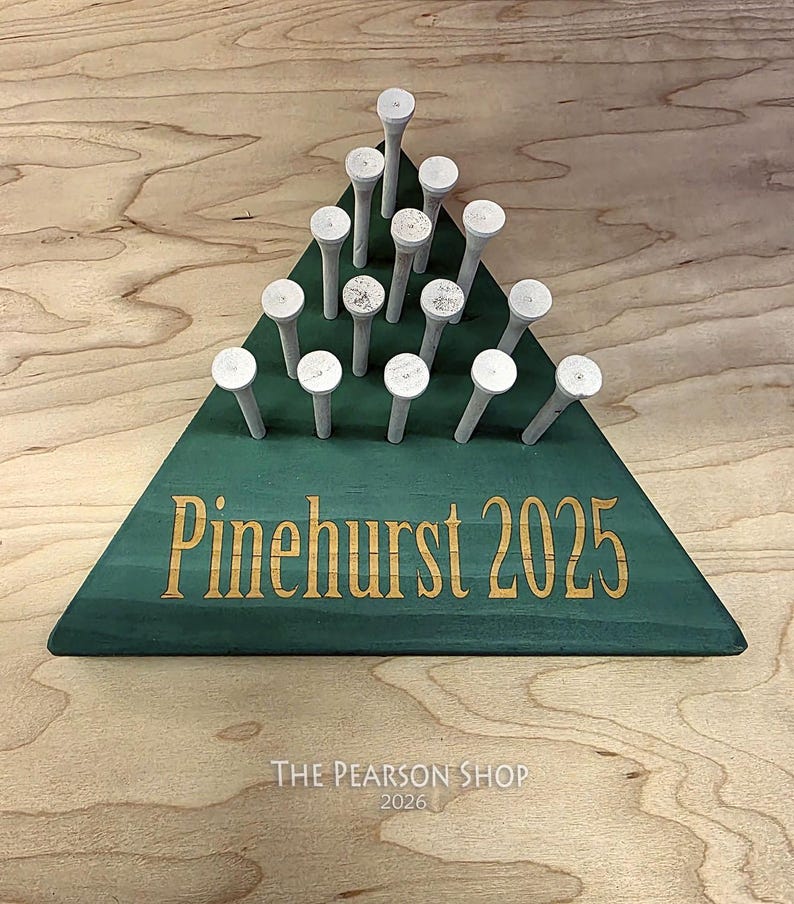 May include: A green wooden golf tee holder in a triangular shape, with the words "Pinehurst 2025" in gold lettering. The holder contains 15 white golf tees. The text "The Pearson Shop 2026" is at the bottom.