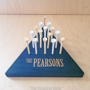 May include: A navy blue wooden triangle with the engraved text "THE PEARSONS" holds golf tees. The tees are a mix of natural wood and white. The base has the text "THE PEARSON SHOP 2025".
