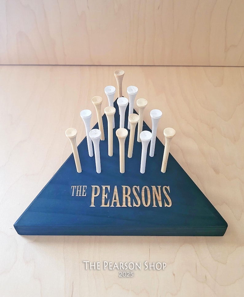 May include: A navy blue wooden triangle with the engraved text "THE PEARSONS" holds golf tees. The tees are a mix of natural wood and white. The base has the text "THE PEARSON SHOP 2025".