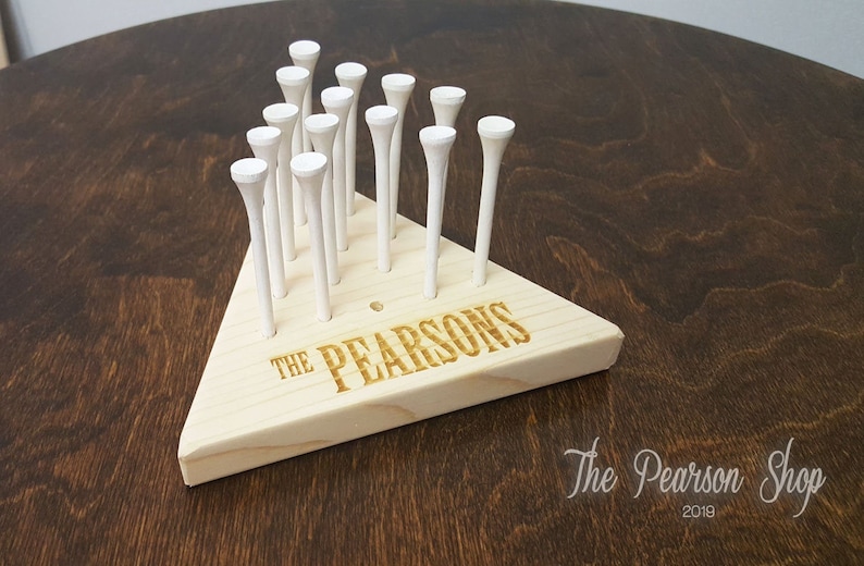 May include: A wooden golf tee holder in a triangular shape, with the text "THE PEARSONS" engraved on the front. The holder contains multiple white golf tees. The background is a dark wood surface.