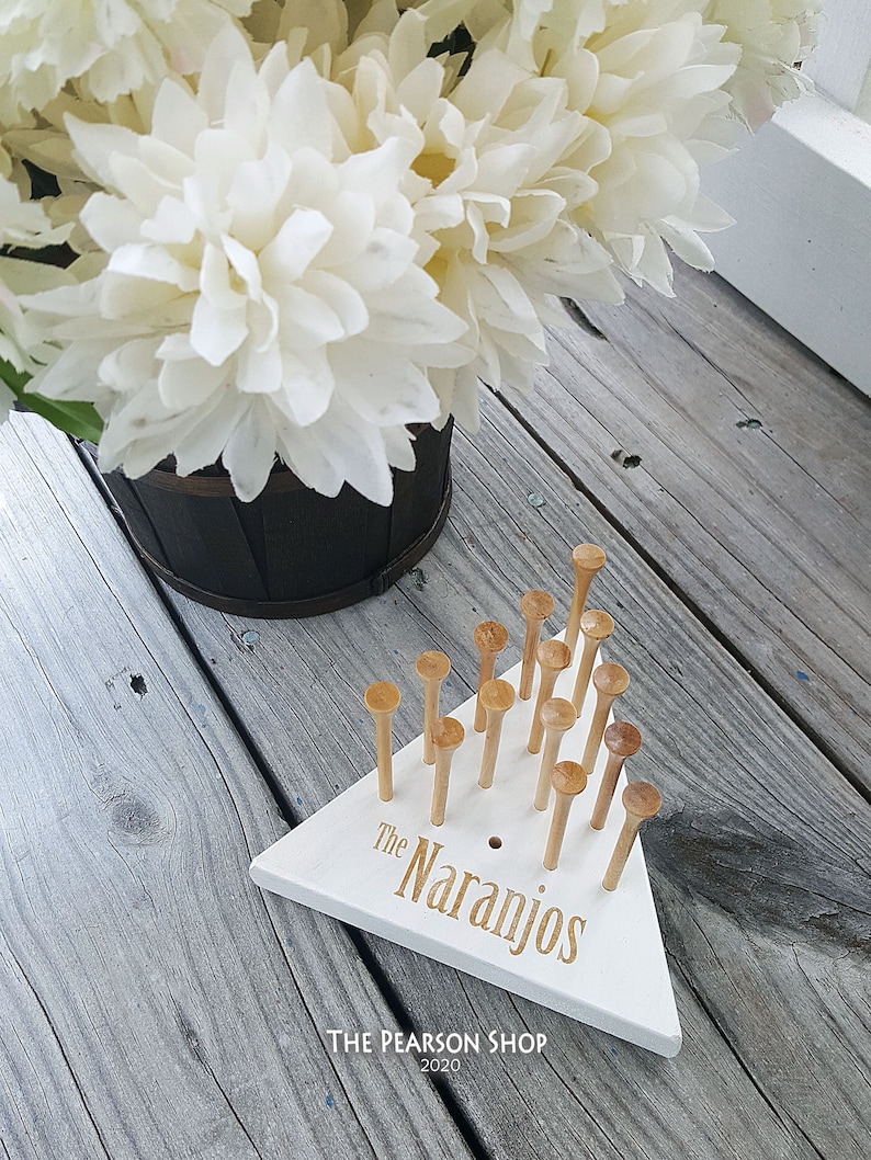 May include: A white wooden peg game with the text "The Naranjos" in gold lettering. The game has wooden pegs and is set on a weathered wooden surface. A bouquet of white flowers is in the background.