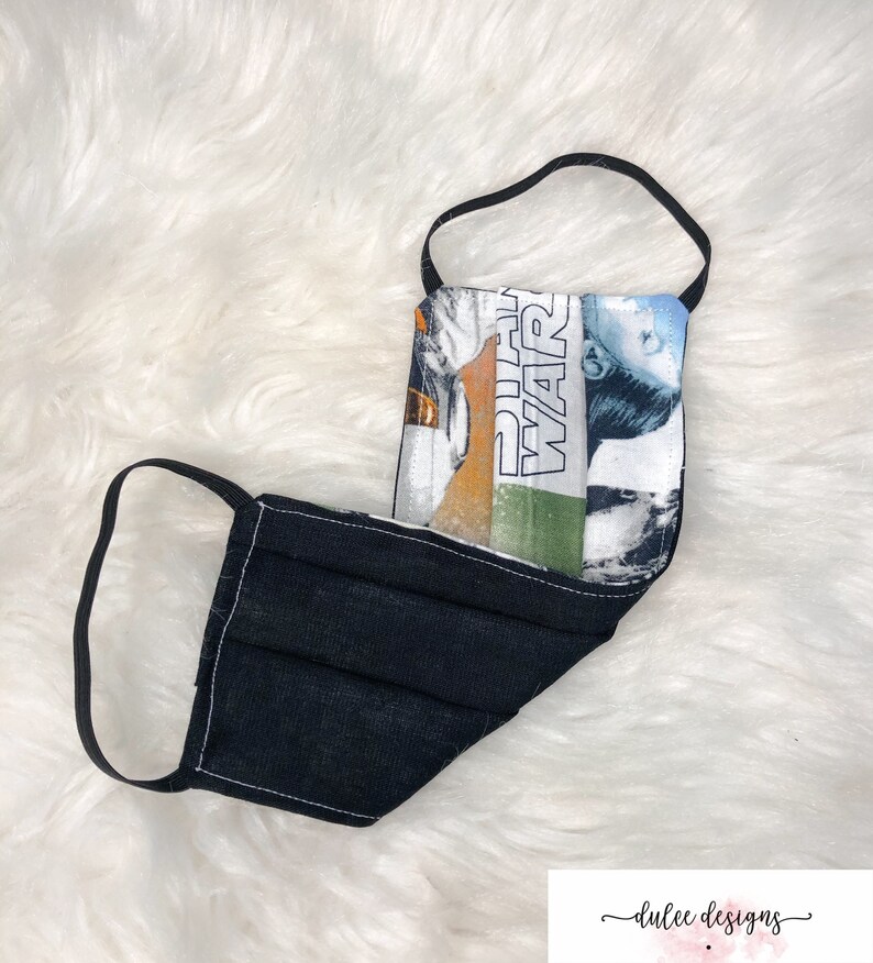 Puede incluir: M&aacute;scara facial reutilizable negra con un panel de tela con tem&aacute;tica de Star Wars que presenta el texto "Star Wars" y un personaje de la pel&iacute;cula.