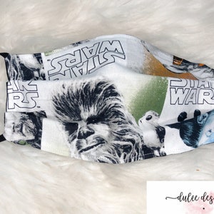 Puede incluir: Una mascarilla facial blanca con una banda el&aacute;stica negra. La mascarilla presenta un estampado de Star Wars colorido con personajes como Chewbacca y Yoda.