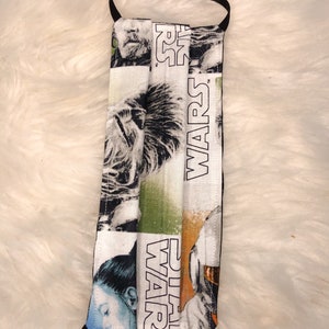 Puede incluir: Una mascarilla facial blanca con una banda el&aacute;stica negra. La mascarilla tiene un tema de Star Wars con im&aacute;genes de personajes de la franquicia de pel&iacute;culas. El texto "STAR WARS" est&aacute; impreso en la mascarilla.