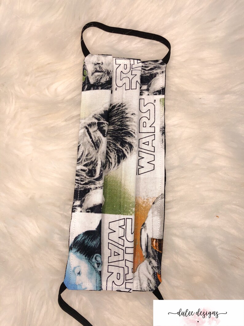 Puede incluir: Una mascarilla facial blanca con una banda el&aacute;stica negra. La mascarilla tiene un tema de Star Wars con im&aacute;genes de personajes de la franquicia de pel&iacute;culas. El texto "STAR WARS" est&aacute; impreso en la mascarilla.