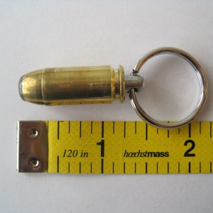 May include: A brass bullet casing keychain with a silver ring. The bullet casing is about 1 inch long and is attached to the ring with a silver swivel. The keychain is lying on a yellow measuring tape with the text "hochstmass GERMANY" printed on it.