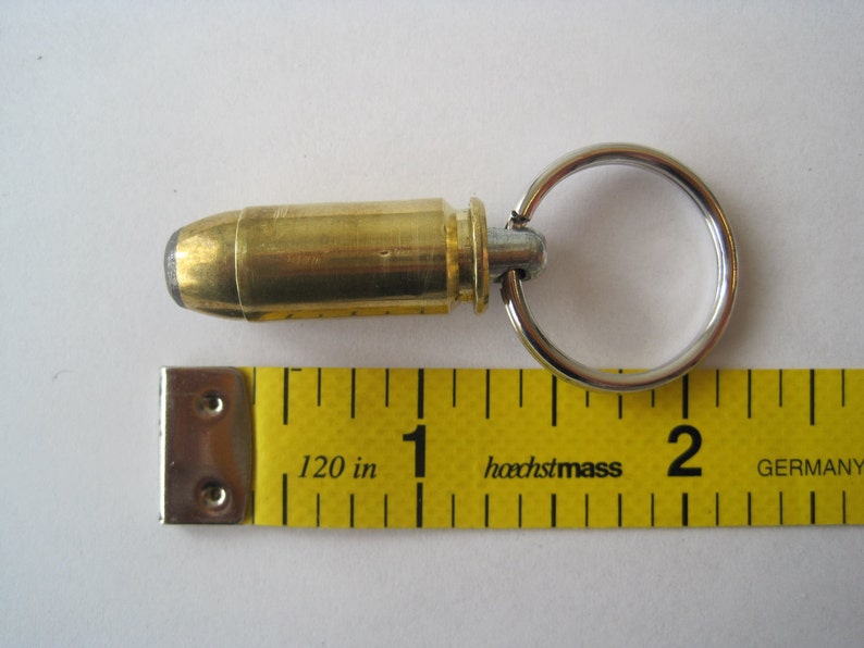 May include: A brass bullet casing keychain with a silver ring. The bullet casing is about 1 inch long and is attached to the ring with a silver swivel. The keychain is lying on a yellow measuring tape with the text "hochstmass GERMANY" printed on it.