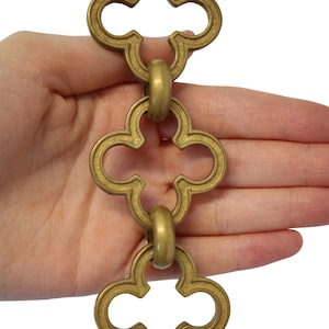 May include: A section of brass-colored decorative chain with a quatrefoil design. The chain links are connected by round rings. The chain is held against a white background.