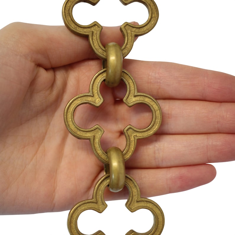 May include: A section of brass-colored decorative chain with a quatrefoil design. The chain links are connected by round rings. The chain is held against a white background.