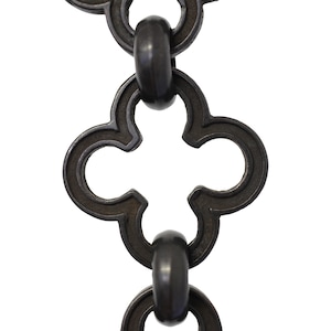 May include: Close-up of a section of a dark metal chain with a repeating quatrefoil design. The chain links are connected by oval rings. The metal has a slightly textured surface and a dark, matte finish. The background is plain white.