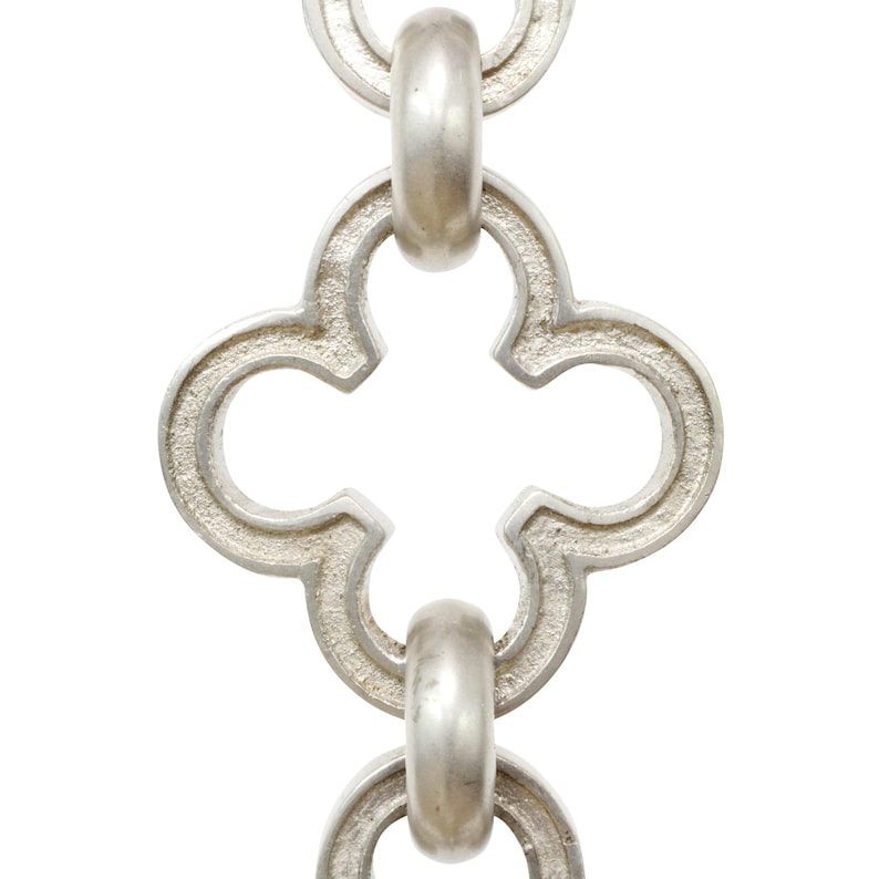 May include: Close-up of a silver-colored chain with a repeating quatrefoil design. The chain links are connected by rounded, oval-shaped connectors. The metal has a slightly textured surface, and the background is white.